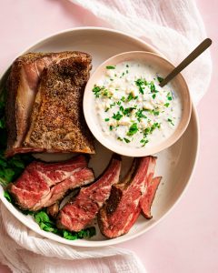 White platter with sliced rack of lamb and bowl of feta sauce on pink surface