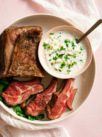 White platter with sliced rack of lamb and bowl of feta sauce on pink surface