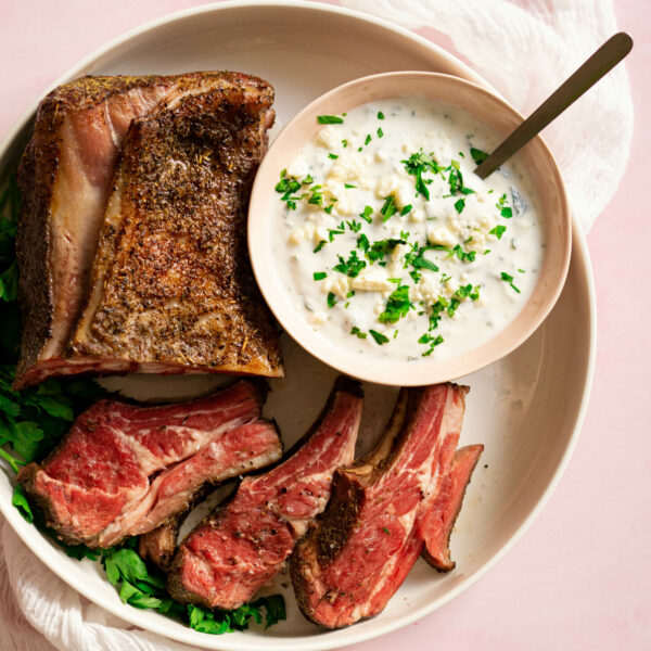 White platter with sliced rack of lamb and bowl of feta sauce on pink surface