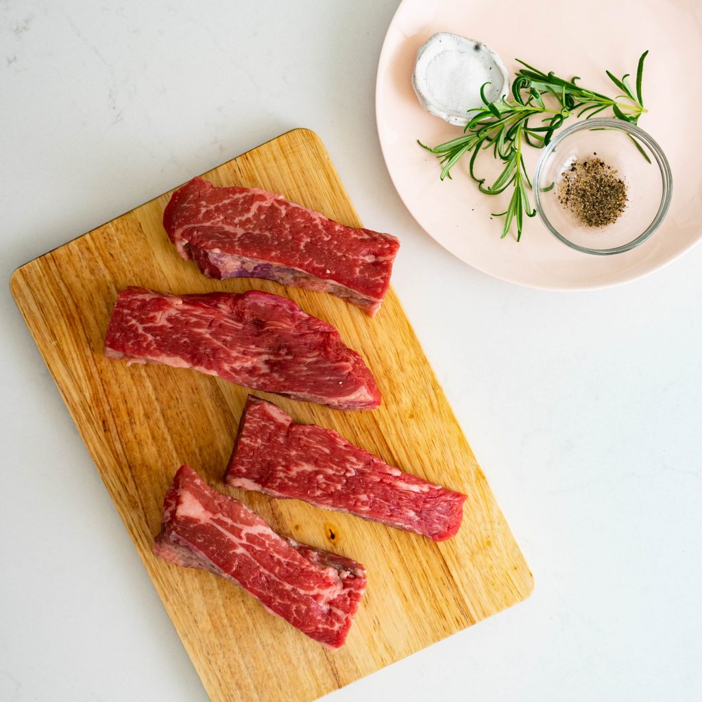 Steak on wood cutting board and salt, pepper, and rosemary on pink plate on white counter