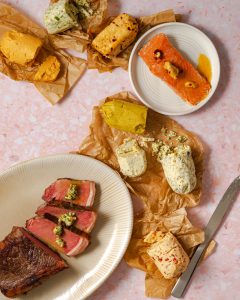 Compound butters on parchment paper, steak on platter, salmon on plate on pink surface