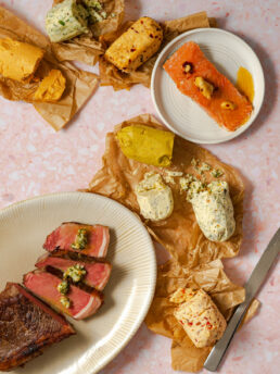Compound butters on parchment paper, steak on platter, salmon on plate on pink surface