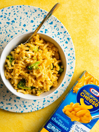 A bowl of macaroni and cheese with broccoli sits on a speckled blue and white plate, alongside a boxed mac and cheese Kraft Macaroni & Cheese box, on a bright yellow background. A spoon rests in the bowl.
