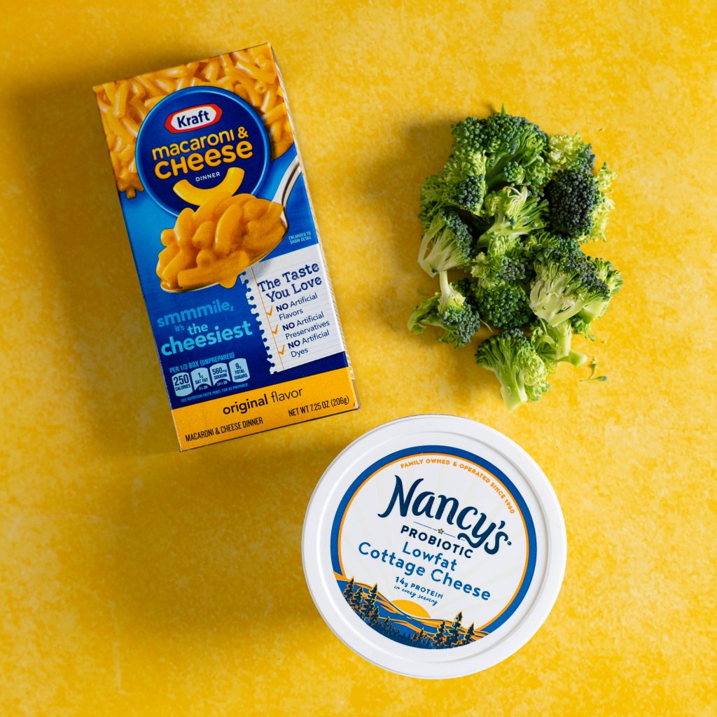 Box of Kraft mac and cheese, container of cottage cheese, and chopped broccoli on a yellow surface
