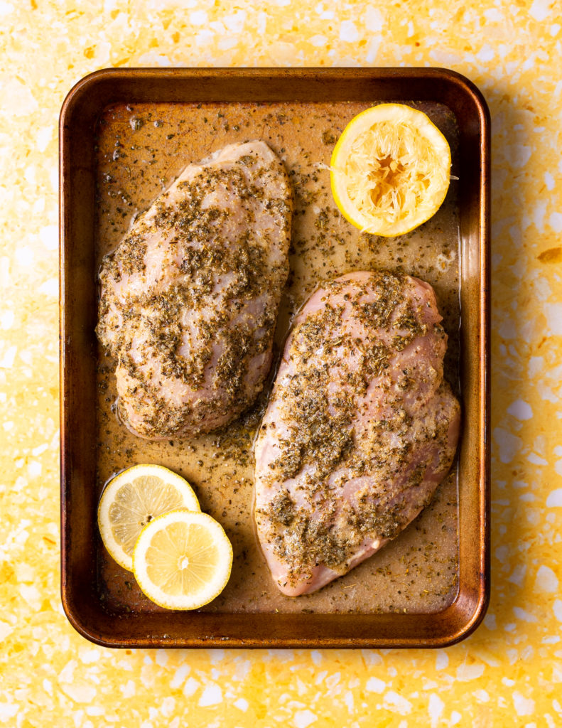Chicken breasts in lemon marinade on sheet pan on yellow terrazzo surface.