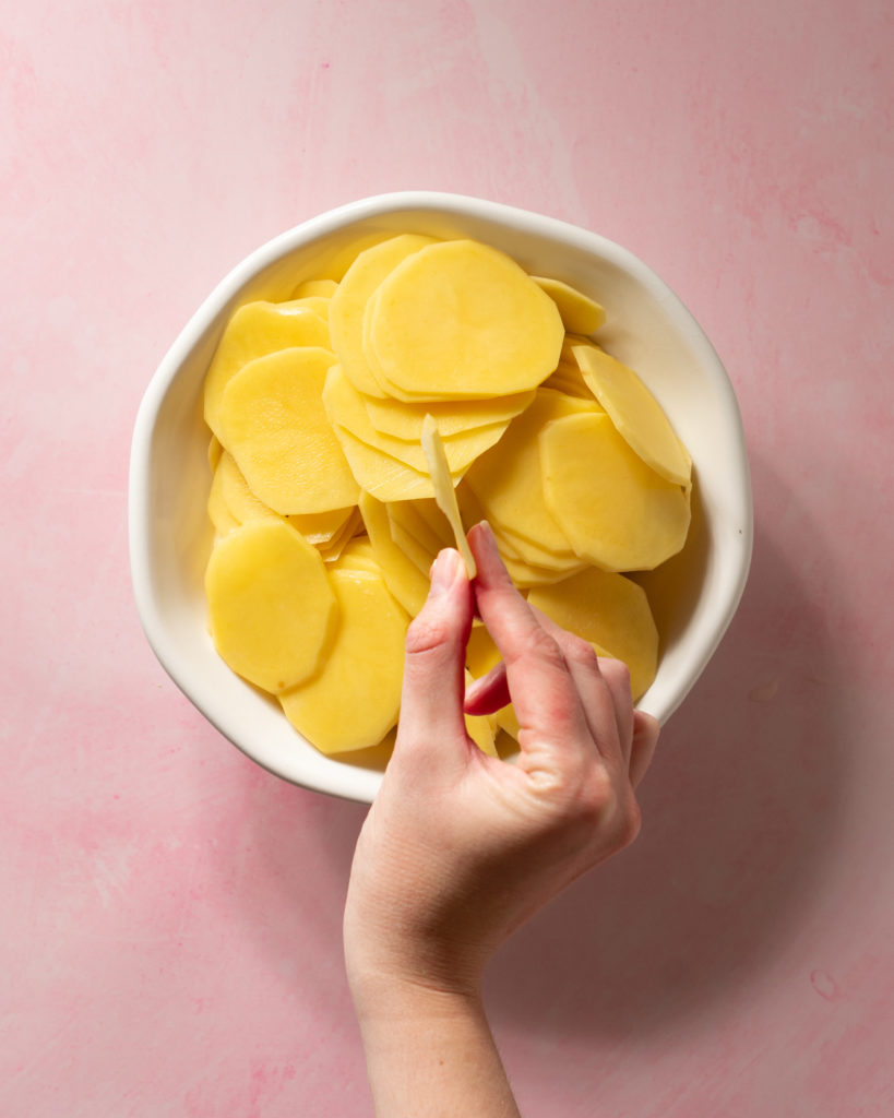 Bowl of slices potatoes on pink surface, hand holding one slice to show how thin potato slices should be for scalloped potatoes