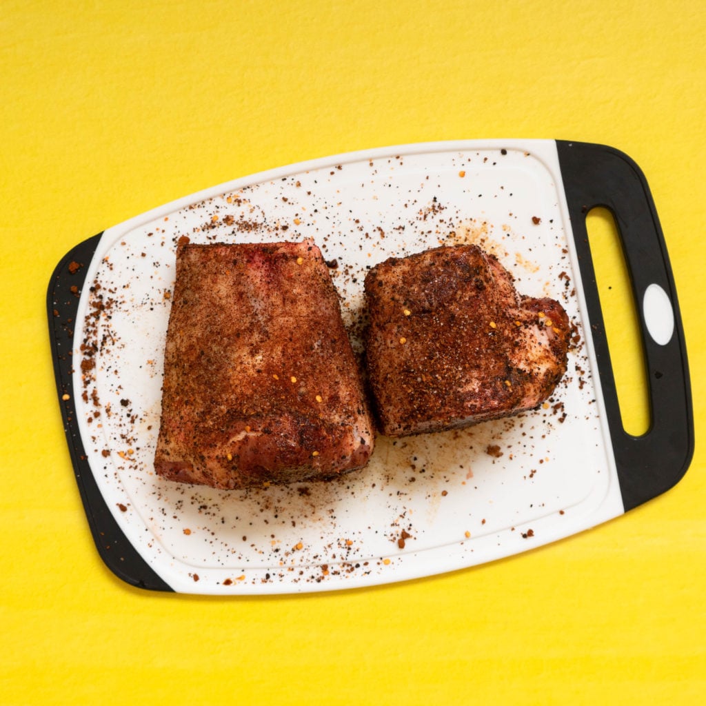 Two pieces of seasoned, uncooked meat sit on a white cutting board with a black handle, against a bright yellow background. Spices are scattered around the meat on the board.