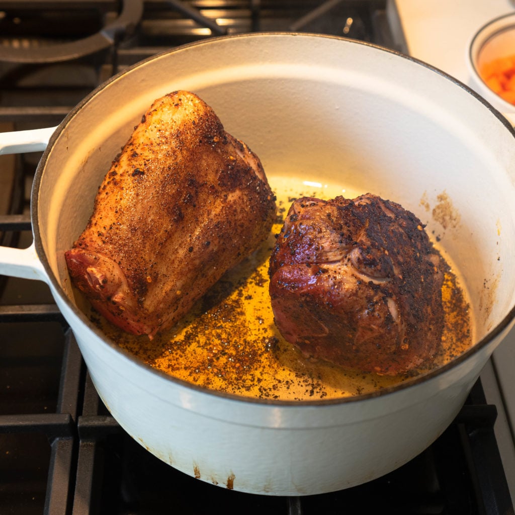 Two seasoned pieces of meat are browning in a white Dutch oven on a stovetop, with spices and oil visible in the pan.