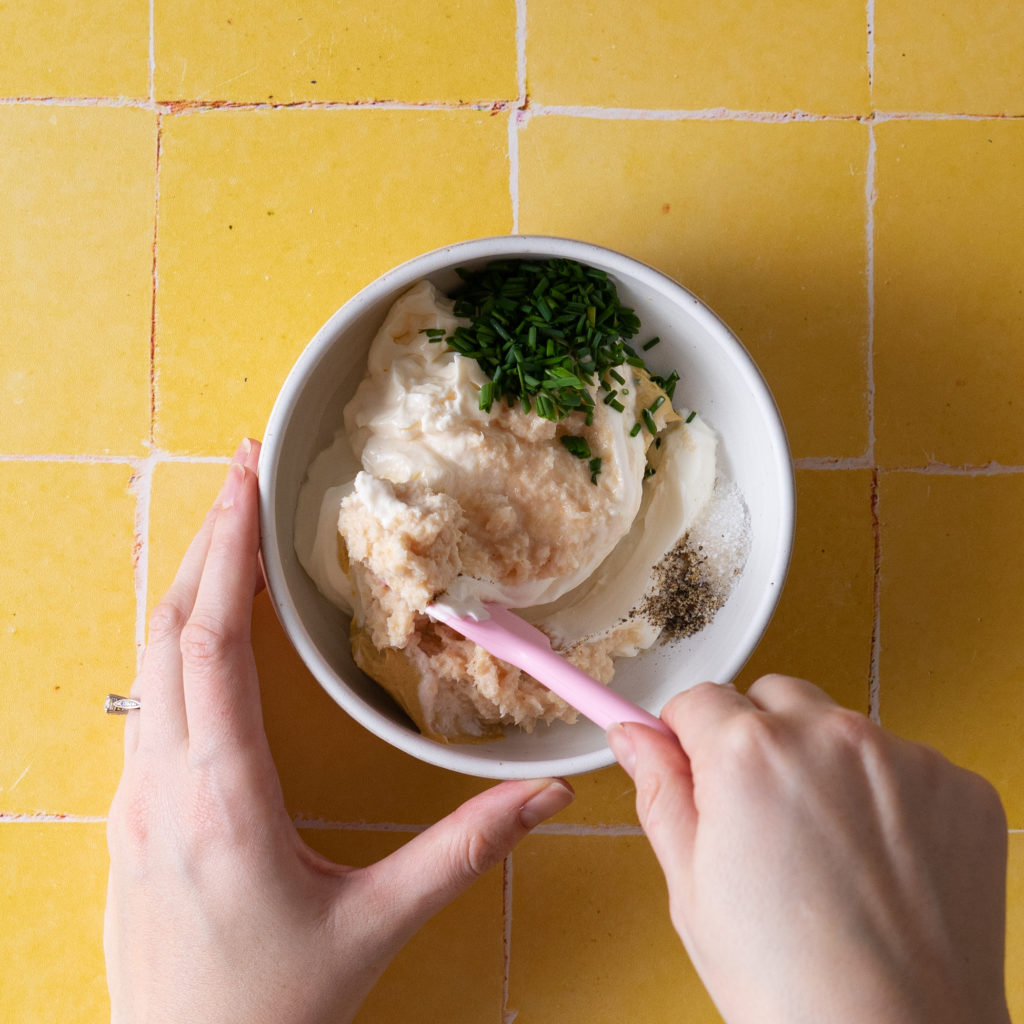 Hand stirring horseradish aioli ingredients in bowl on yellow tile.