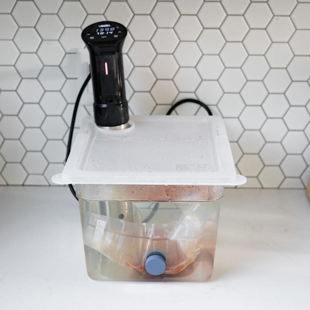 Vacuum sealed pork brisket in sous vide water bath on white counter top.