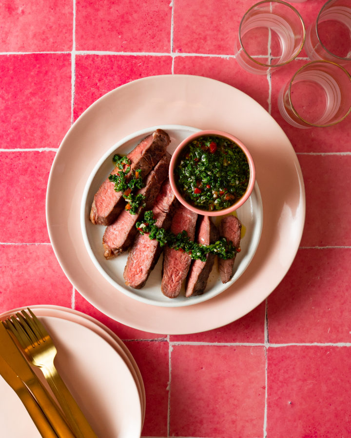 Steak on a white plate drizzled with chimichurri next to a bowl of chimichurri on bright pink tile.