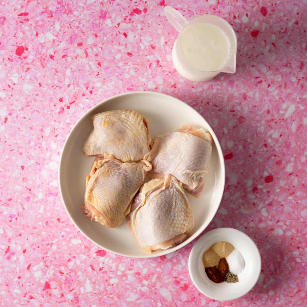Raw chicken thighs on a plate, buttermilk in a pitcher, spices in a small white bowl on a pink surface