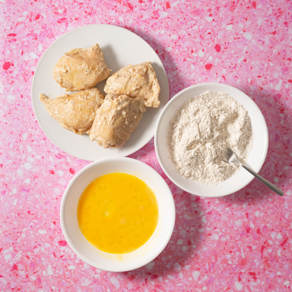 Shallow bowl with eggs, shallow bowl with seasoned flour, and cooked chicken thighs on pink background