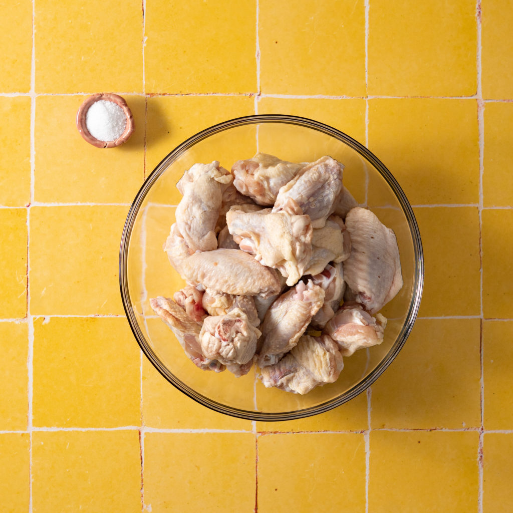 Bowl of raw chicken wings and pinch bowl with salt on yellow tiled surface.