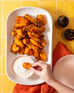 Buffalo wings on white platter, hand dipping wing into blue cheese sauce on yellow background.
