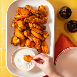 Buffalo wings on white platter, hand dipping wing into blue cheese sauce on yellow background.