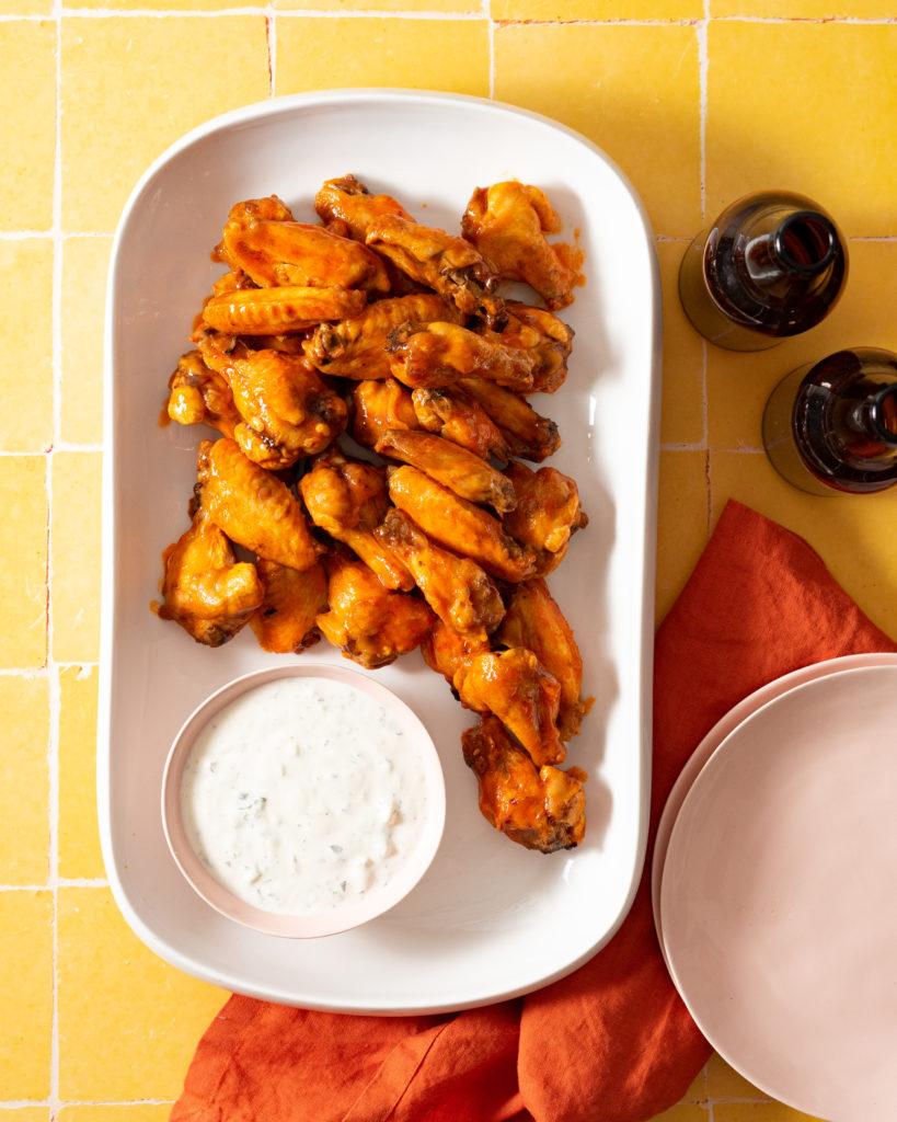 Buffalo wings and blue cheese sauce on white platter on yellow tiled surface.