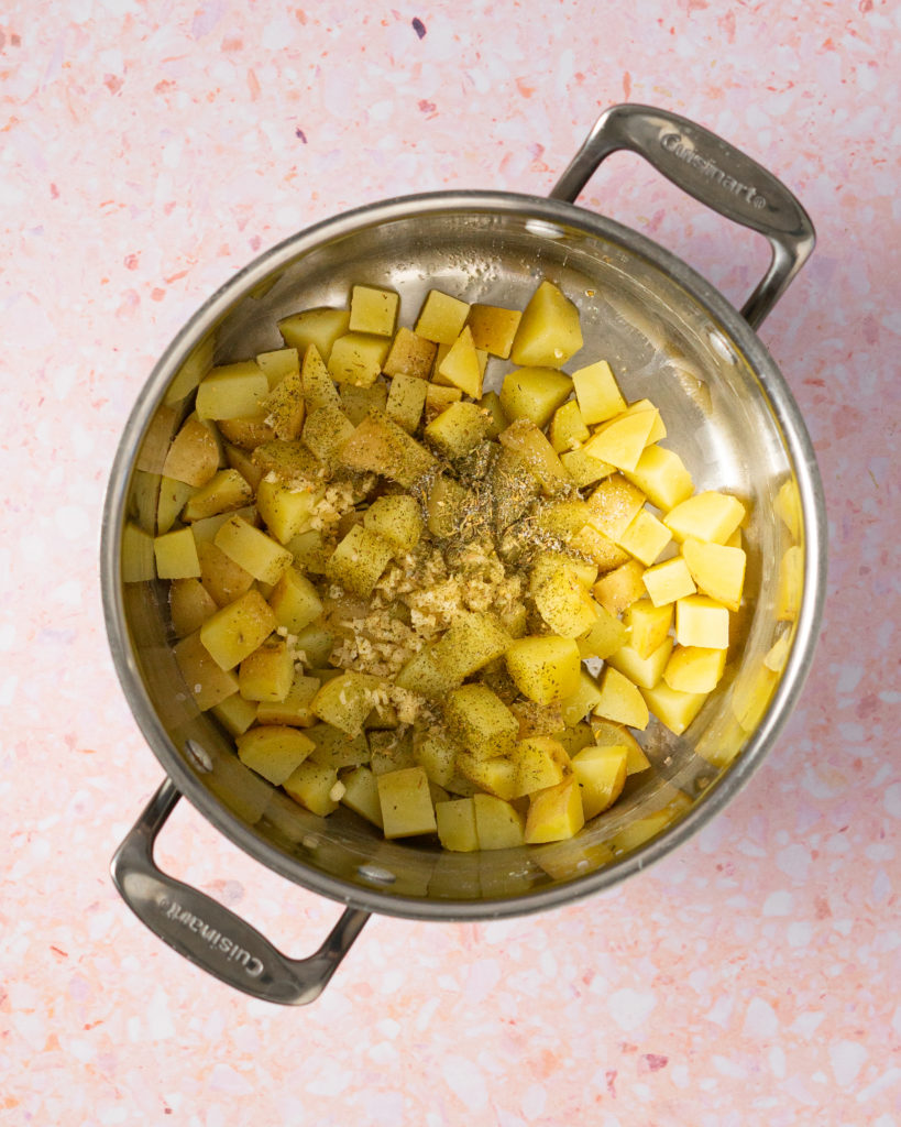 Cooked potatoes, seasoning, garlic in stockpot on pink surface.