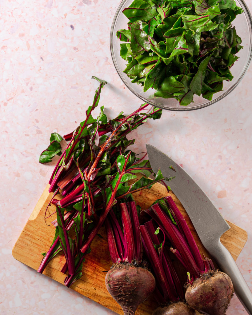 Beet greens in a bowl, beets and stems on wood cutting board on pink surface.