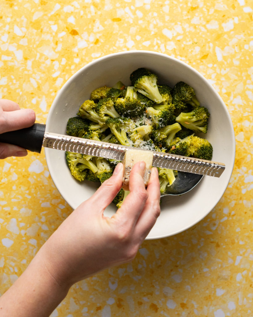 Grating parmesan over the broccoli in a bowl.
