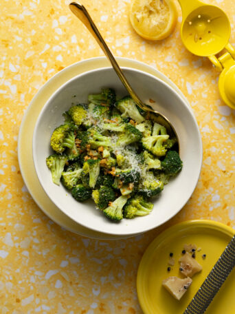 Sous vide broccoli in white bowl on yellow background with parmesan and lemon.