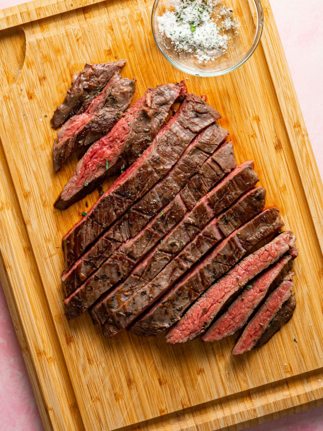 Sliced sous vide flank steak on a wood cutting board with a bowl of herby salt.