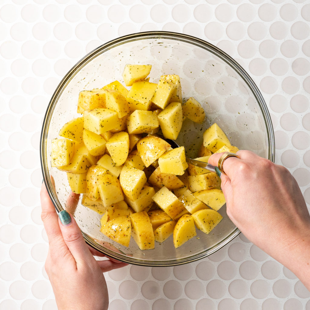 Diced yellow potatoes in bowl with olive oil and seasonings, hands stirring them together