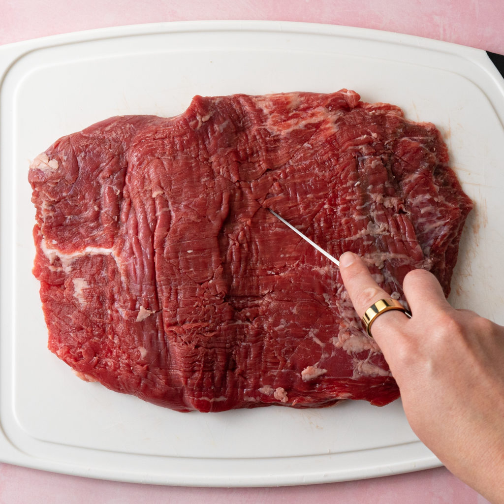 Flank steak on white cutting board being scored with knife.