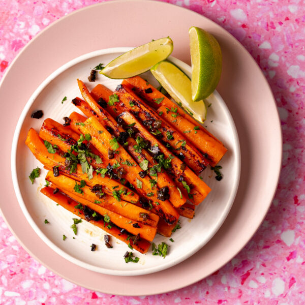 Sous vide carrots on a plate with jalapenos, cilantro, and limes on a pink surface.