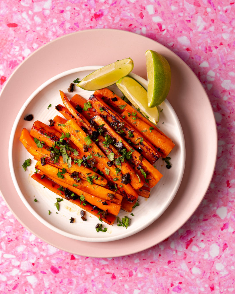 Sous vide carrots on a plate with jalapenos, cilantro, and limes on a pink surface.