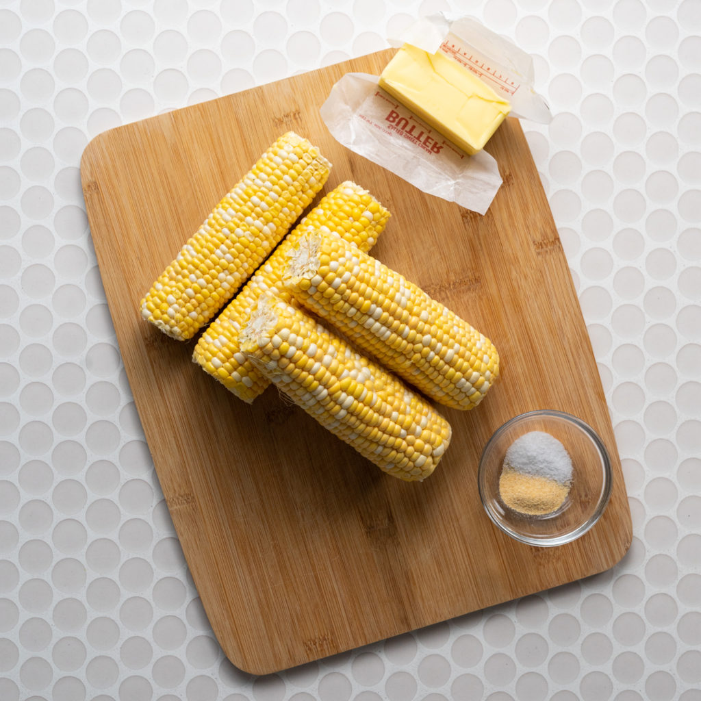 Corn cobs, cube of butter, salt, and garlic powder on wood cutting board