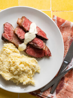Sliced sous vide chuck roast with horseradish sauce and mashed potatoes on a white plate on yellow surface