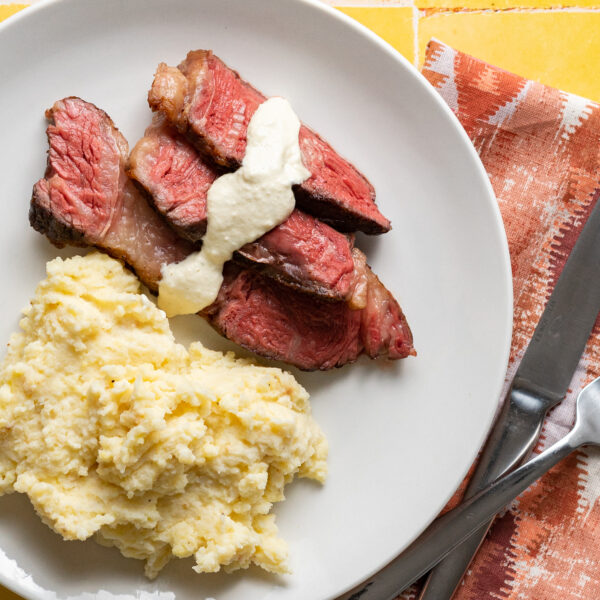Sliced sous vide chuck roast with horseradish sauce and mashed potatoes on a white plate on yellow surface