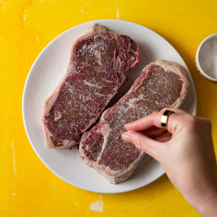 Steak being seasoned with salt on white plate.