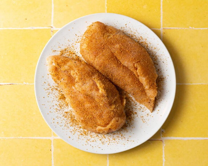 Two raw sous vide chicken breasts coated with a brown spice rub are placed on a white plate, which sits on a yellow tiled surface.