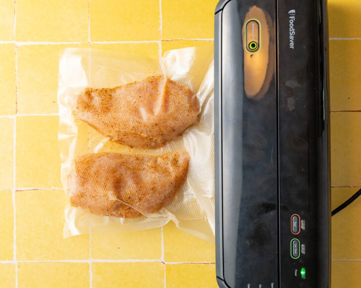 Two seasoned sous vide chicken breasts are being vacuum-sealed in a plastic bag using a FoodSaver vacuum sealer on a yellow tiled countertop.