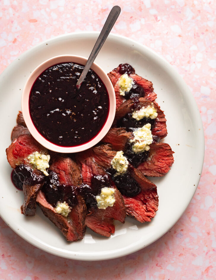 Sliced elk roast on a plate with blue cheese butter and blueberry sauce.