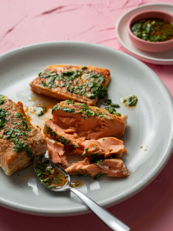 A plate with sous vide salmon fillets topped with green herb sauce. One fillet is flaked with a spoon, revealing the tender pink interior. A small bowl of extra herb sauce sits in the background on a pink surface.