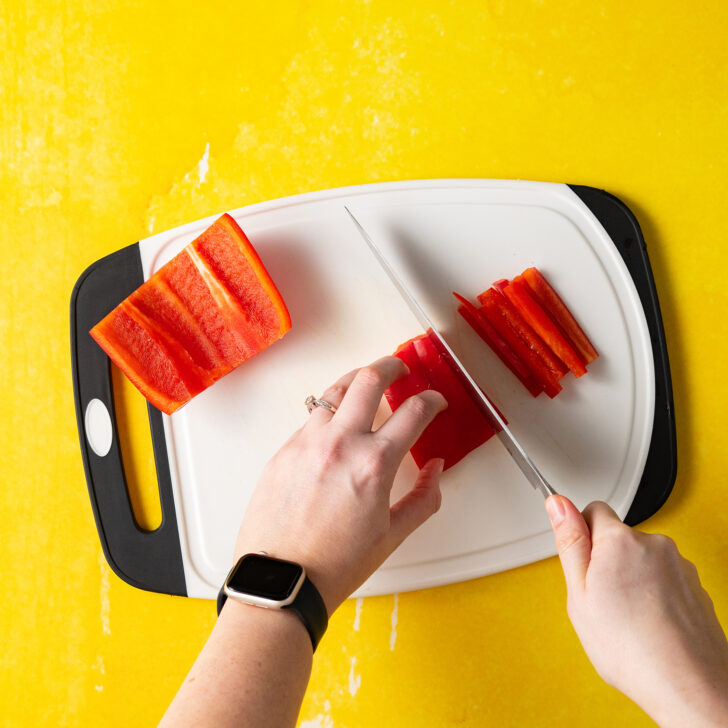 Slicing red bell pepper into thin strips on white cutting board.