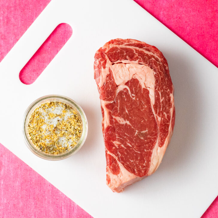 Ribeye steak on white cutting board with bowl of seasoning.