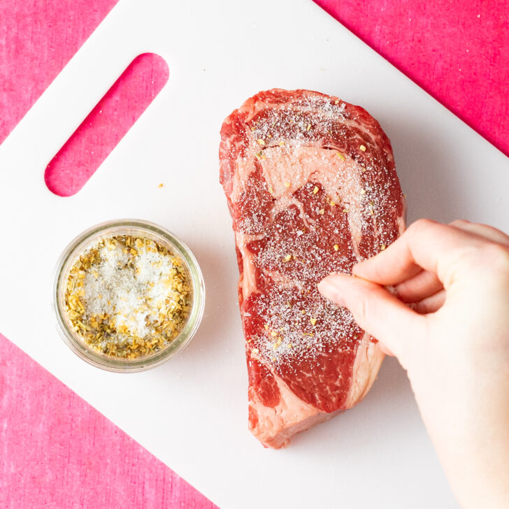 Hand seasoning ribeye steak on white cutting board.