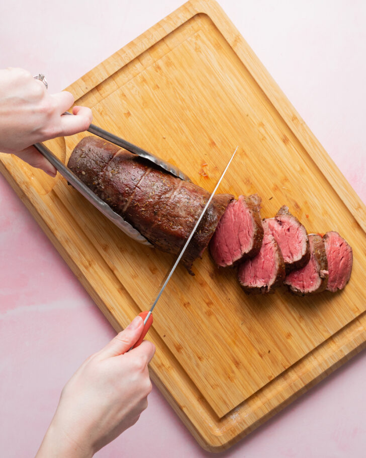 Beef tenderloin on wood cutting board being sliced into rounds.