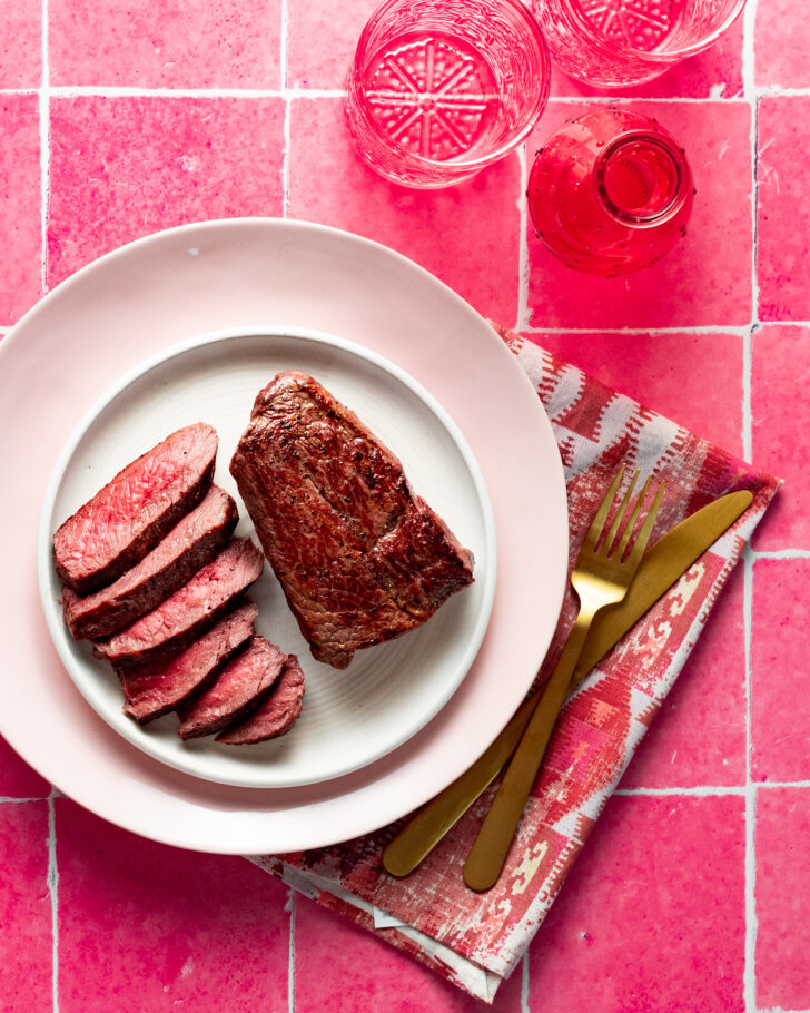 Sliced sous vide medium-rare sirloin steak on a plate with a whole steak on a pink background.