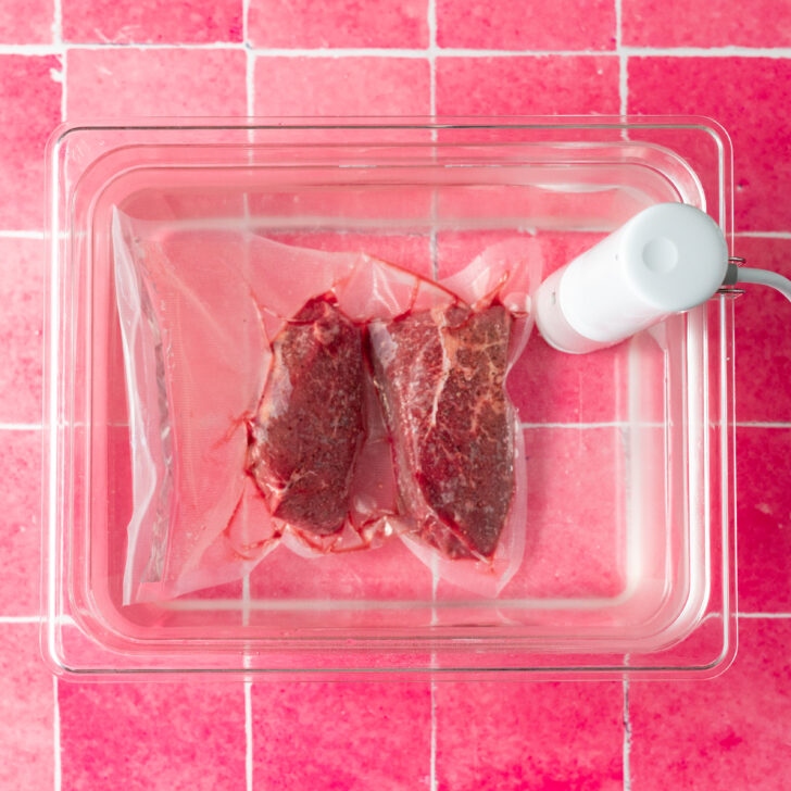 Vacuum sealed steaks in sous vide water bath on pink surface.