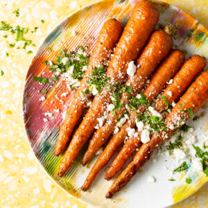 Smoked carrots with za'atar, honey, feta, and parsley on a colorful plate on a yellow backdrop.