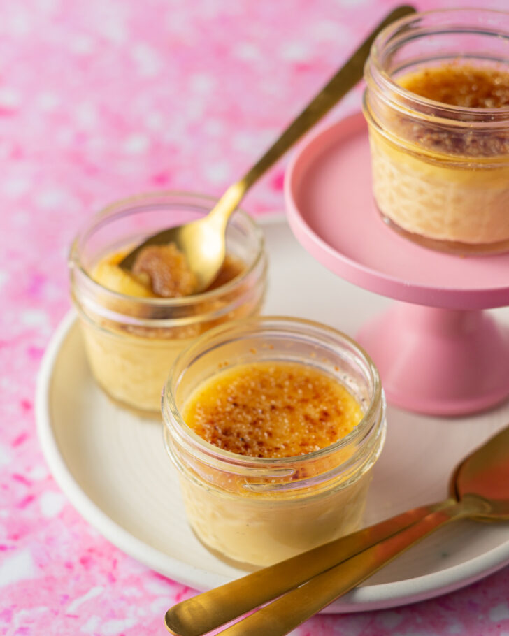 Sous vide crème brûlée in jars on white plate on pink surface with gold spoons.