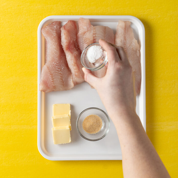 Seasoning cod with salt on white tray on yellow background.