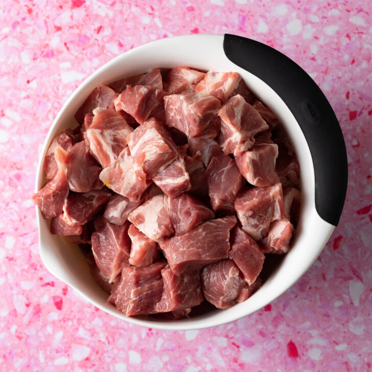 White bowl filled with cubed pork shoulder on pink surface.