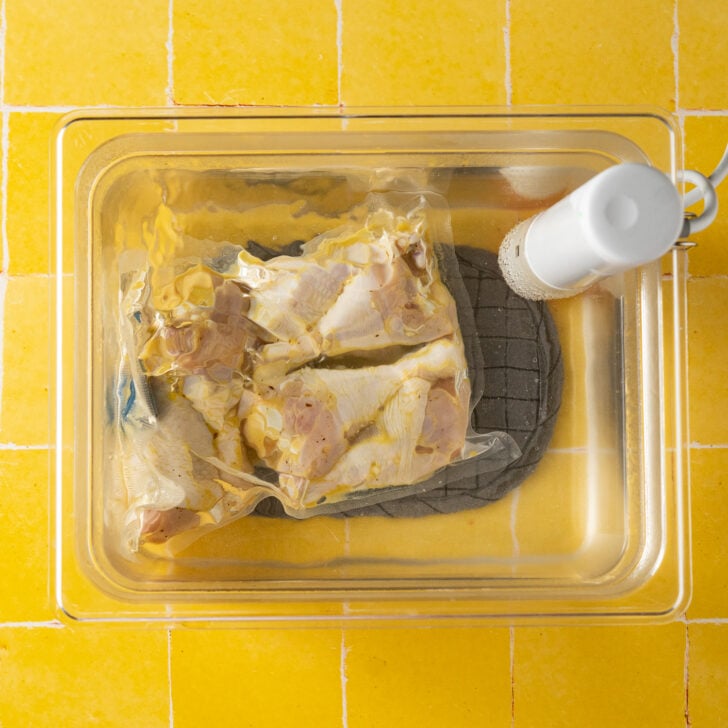 Vacuum-sealed sous vide chicken legs in a plastic bag are submerged in a clear water container with an immersion circulator, on a yellow tiled surface.