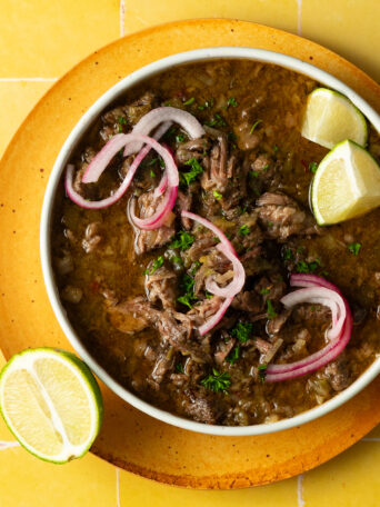 Sous vide barbacoa served in a bowl with red onions and limes on a yellow surface.